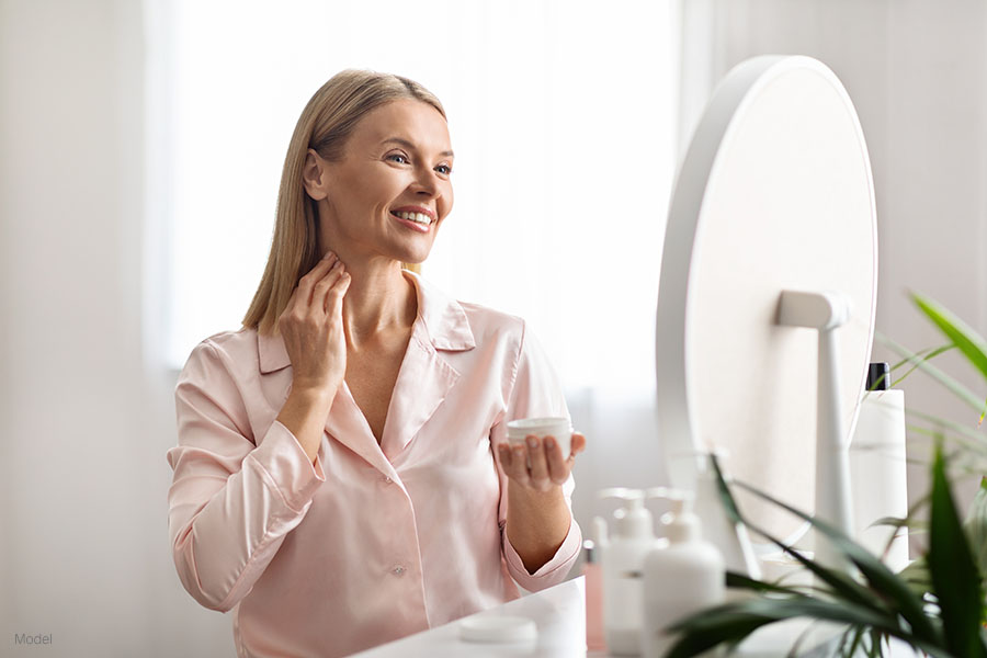 Beautiful middle aged woman smiling in a mirror while she applies moisturizing cream to her neck